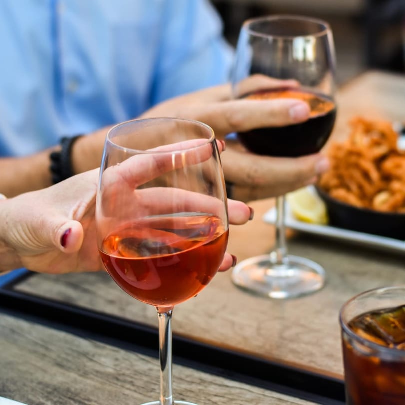Two people holding glasses of red and rosé wine, with a dish of fried food in the background.