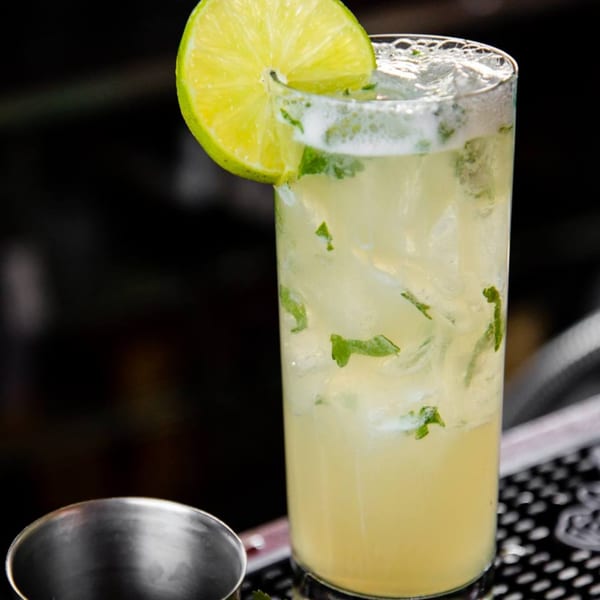 A tall glass of mojito with ice, mint leaves, and a lime wedge on a dark bar.