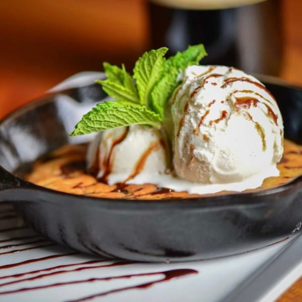 Warm chocolate chip cookie skillet with two scoops of vanilla ice cream, chocolate drizzle, and mint.