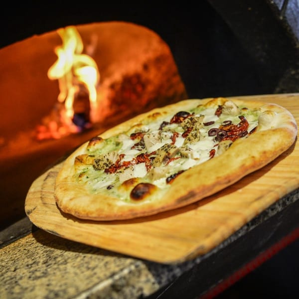 A freshly baked flatbread pizza with chicken, sun-dried tomatoes, and olives, on a wooden peel near a fiery oven.