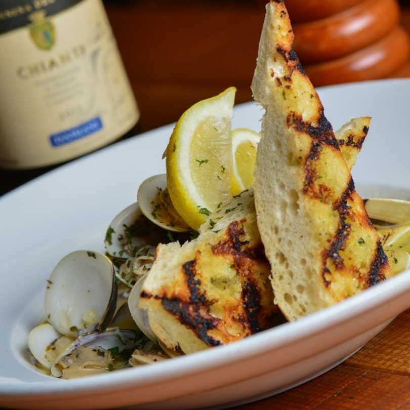 Clams in a white bowl with lemon wedges, grilled bread, and a bottle of Chianti in the background.