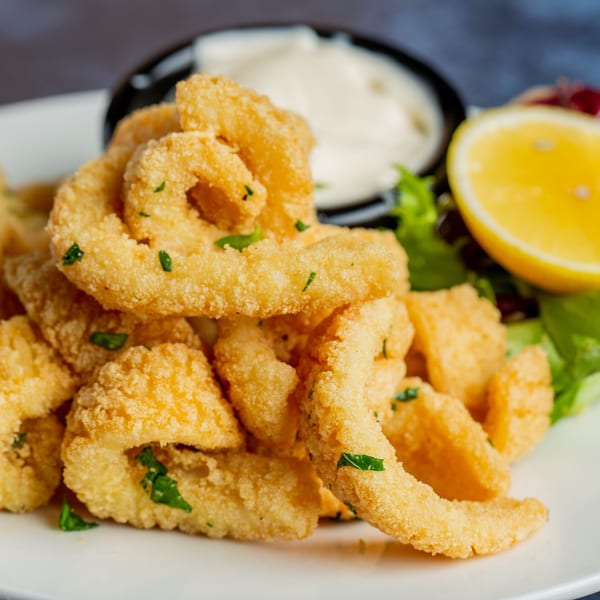 Crispy fried calamari rings with a side of white dipping sauce, lemon wedge, and lettuce.