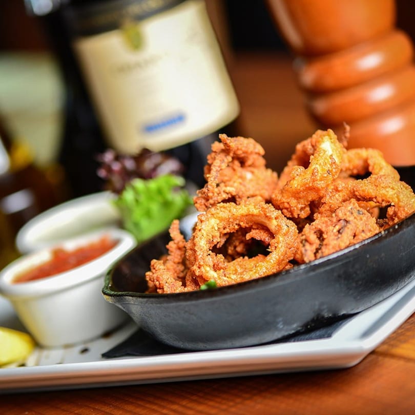 Crispy fried calamari in a black bowl with dipping sauces and a wine bottle in the background.