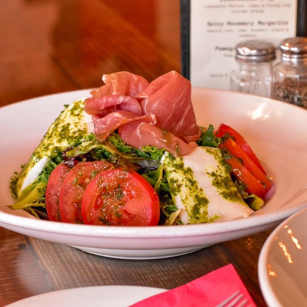 A white bowl filled with a caprese salad, prosciutto, tomatoes, mozzarella, and pesto.