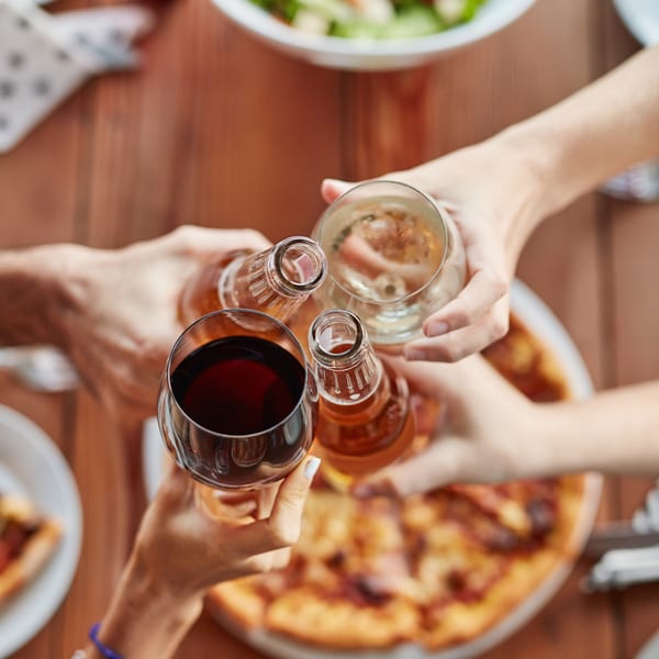 Four people clinking glasses and bottles of drinks over a pizza and salad on a wooden table.