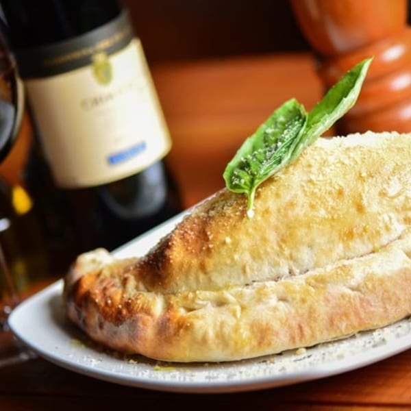 Golden-brown calzone topped with parmesan and basil, next to a bottle of red wine.