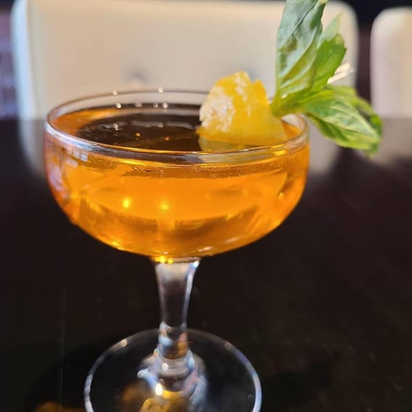 A vibrant orange cocktail in a coupe glass, garnished with a lemon peel and fresh basil.