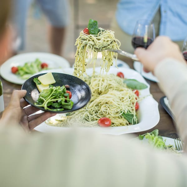 Hands serving pesto pasta with cherry tomatoes and basil, alongside a small plate of greens and cheese.