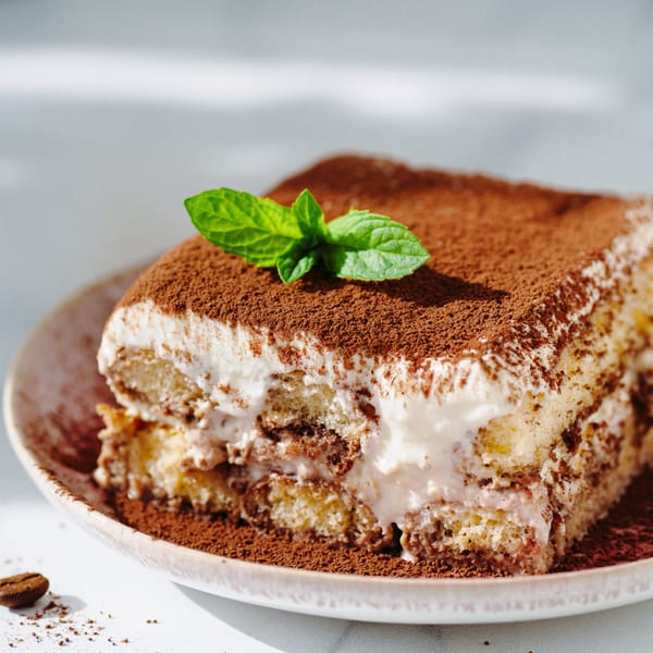 A slice of tiramisu with cocoa powder and a mint leaf on a light plate.