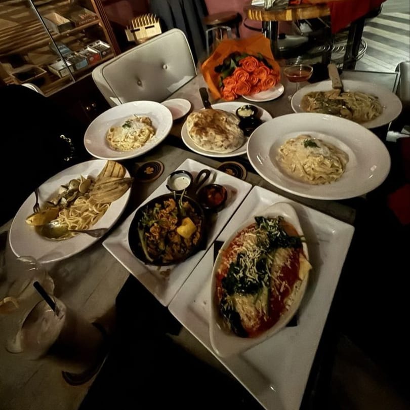 Table laden with various pasta dishes, a pan of food, and a bouquet of red roses.