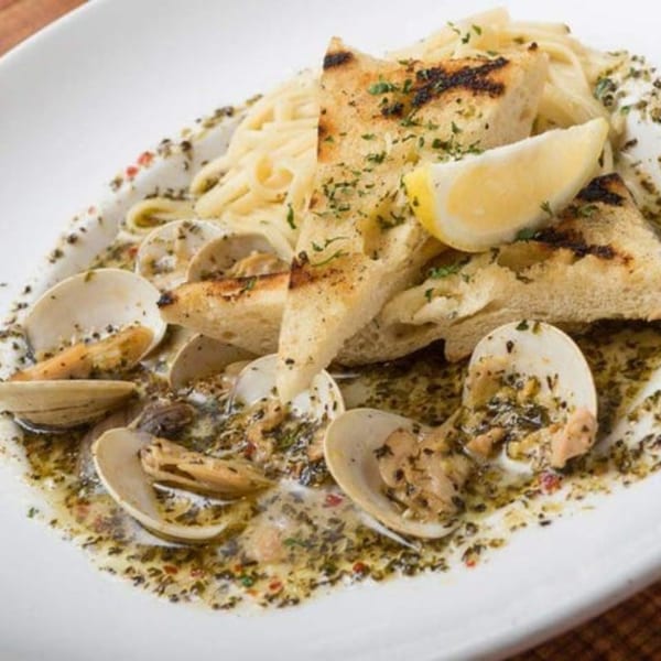 Clams and pasta in a green sauce, with grilled bread and a lemon wedge on a white plate.